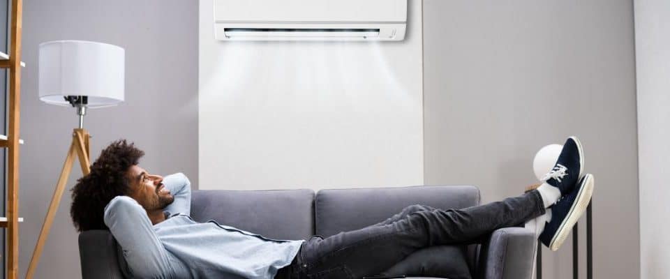Un homme se détend sur un canapé sous un climatiseur mural, avec une lampe et une étagère en arrière-plan.